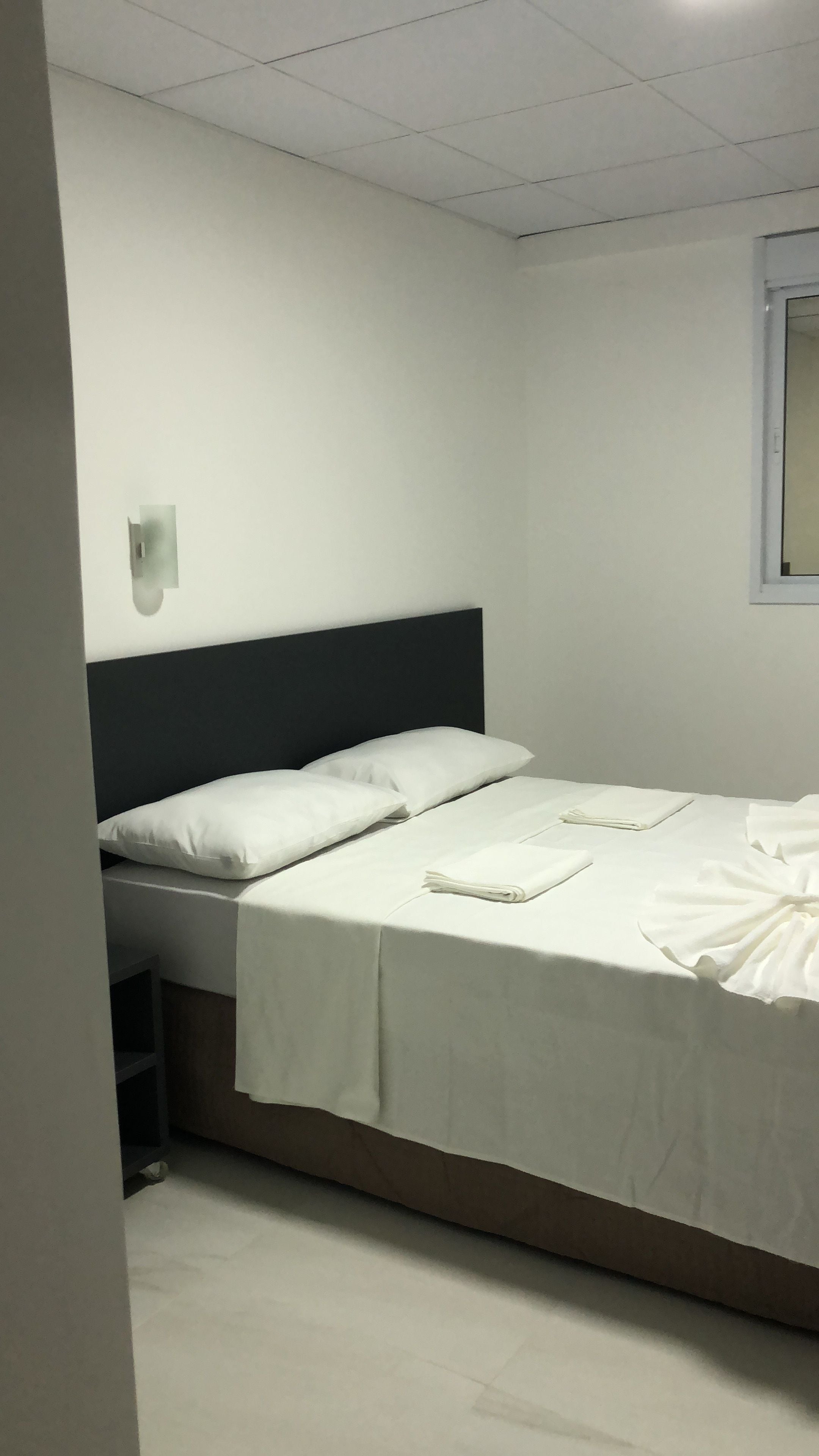A neatly made bed with white linens in a room with white walls and a tiled floor.