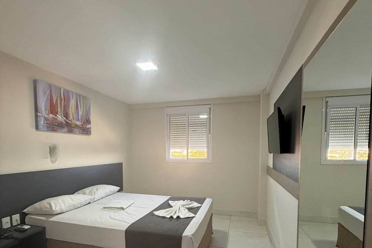 Un dormitorio con una cama, una mesita de noche, un televisor, una ventana con persianas y un cuadro en la pared.