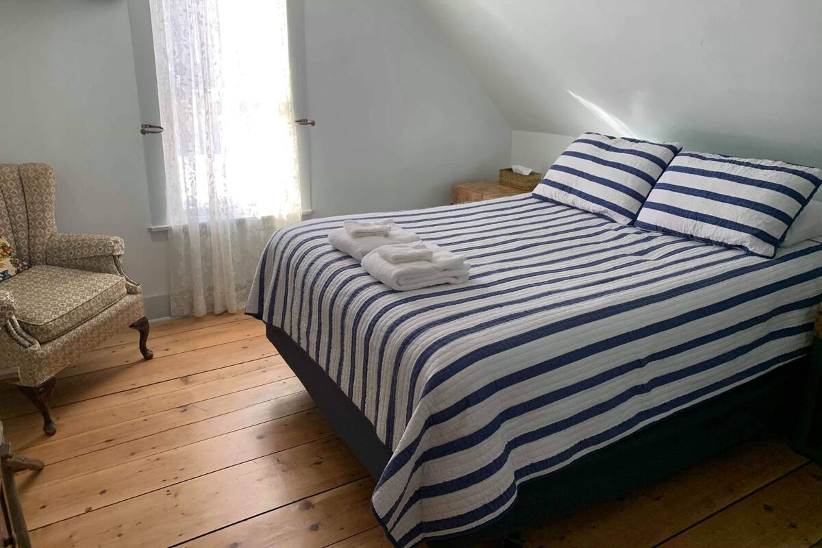 A bedroom with a bed, a chair, a window, and a bedside table.