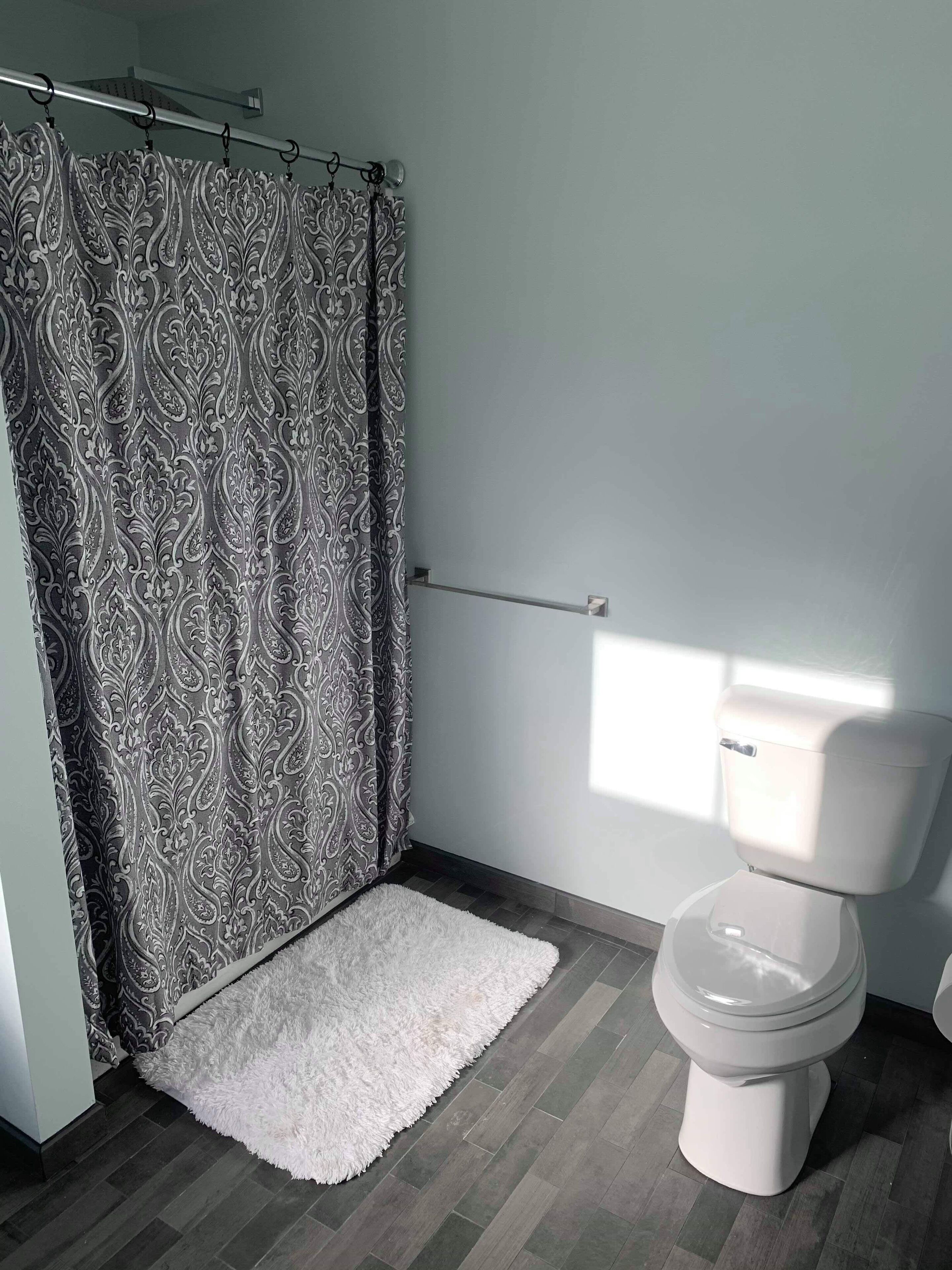 Un baño con cortina de ducha estampada, un felpudo blanco mullido, un inodoro y un perchero para toallas.