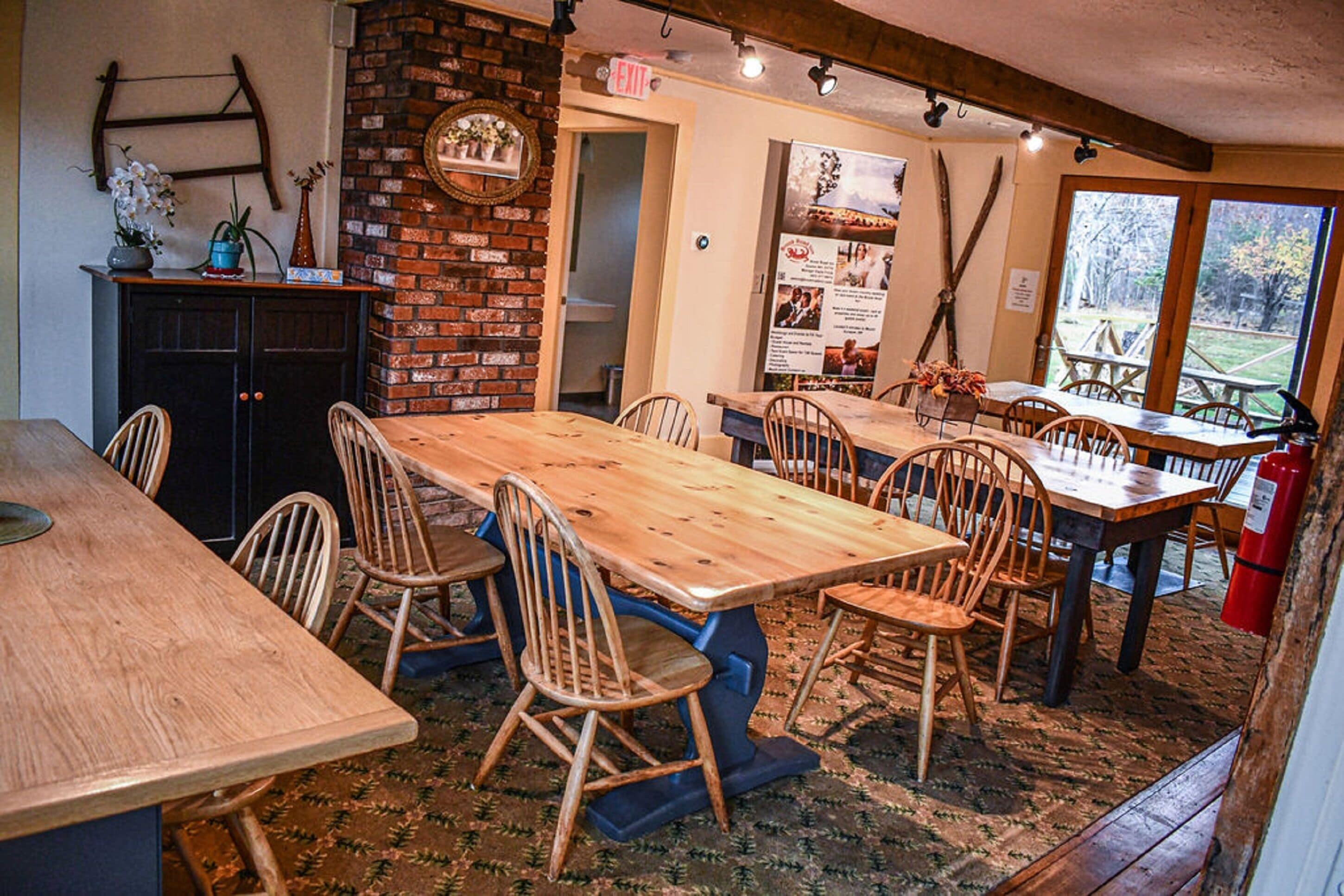 A rustic dining area with a long wooden table and chairs, a brick wall, and a view of the outdoors through large windows.