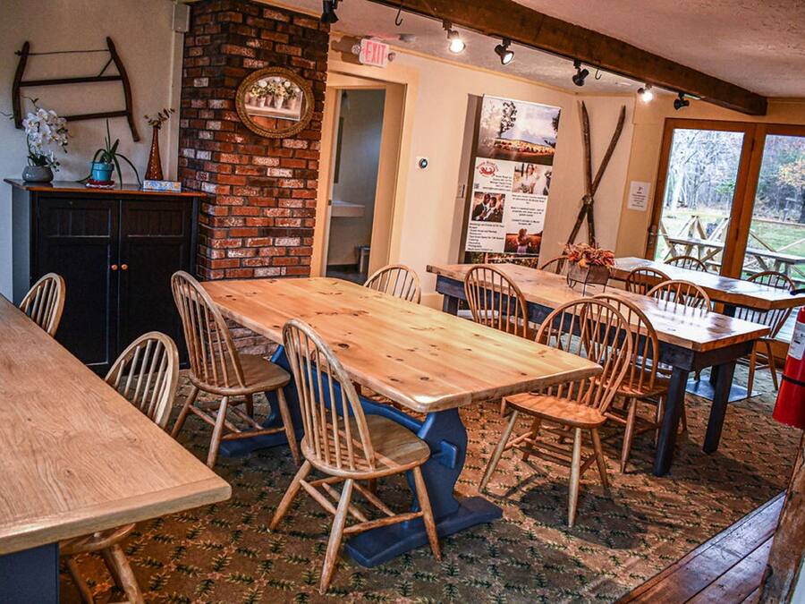 A rustic dining area with a long wooden table and chairs, a brick wall, and a view of the outdoors through large windows.