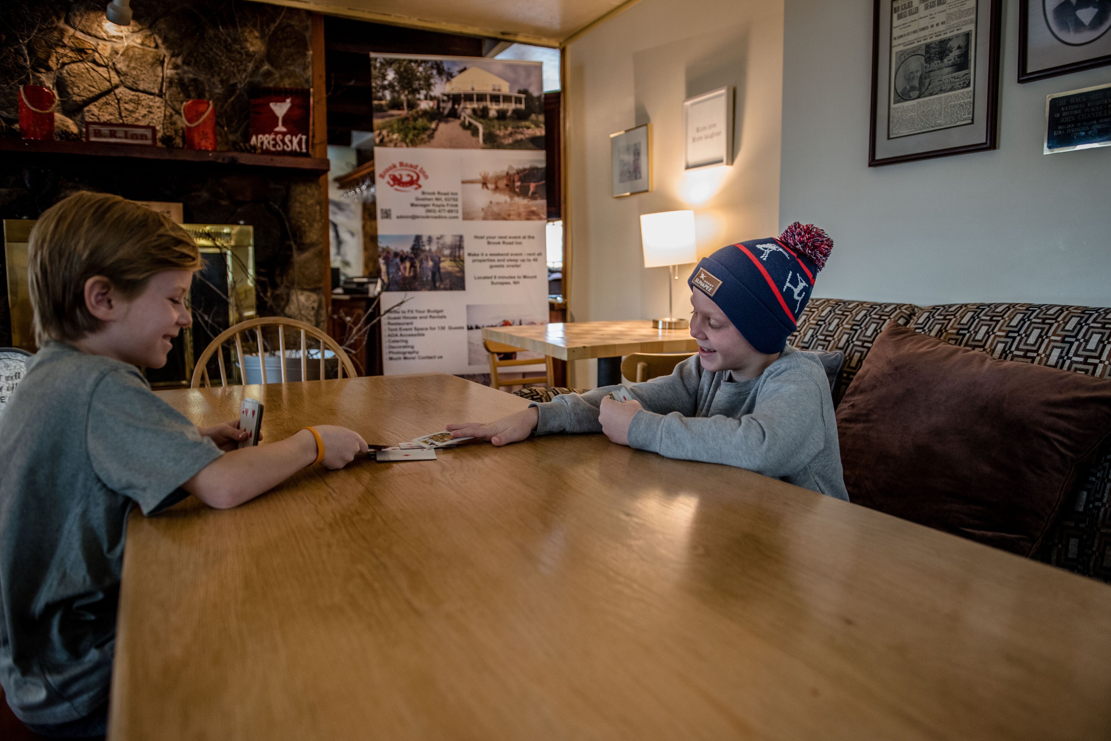 Two people playing cards at a wooden table in a room with a stone wall, a lamp, and framed pictures.
