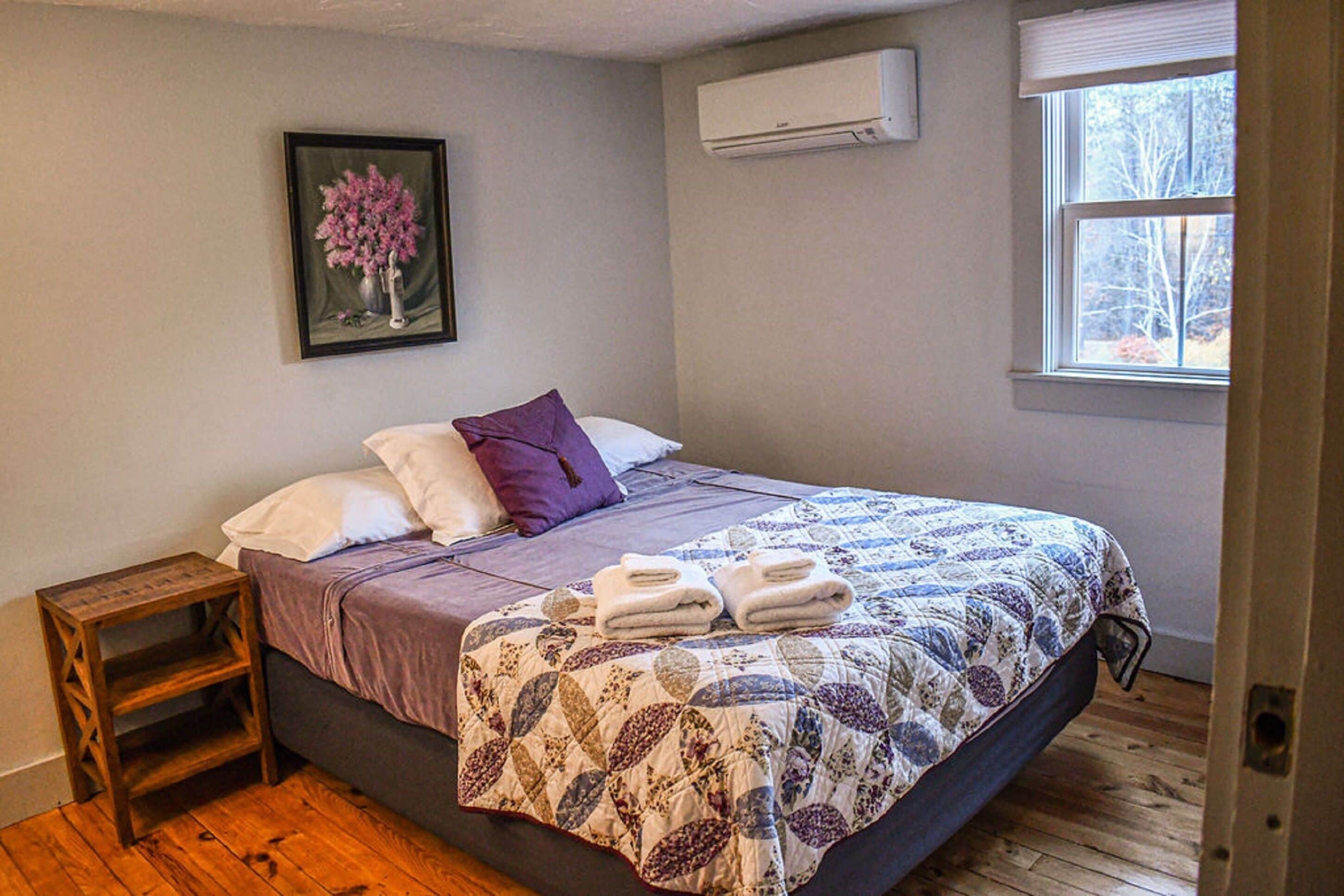 A bedroom with a bed, a nightstand, a painting, an air conditioner, and a window.
