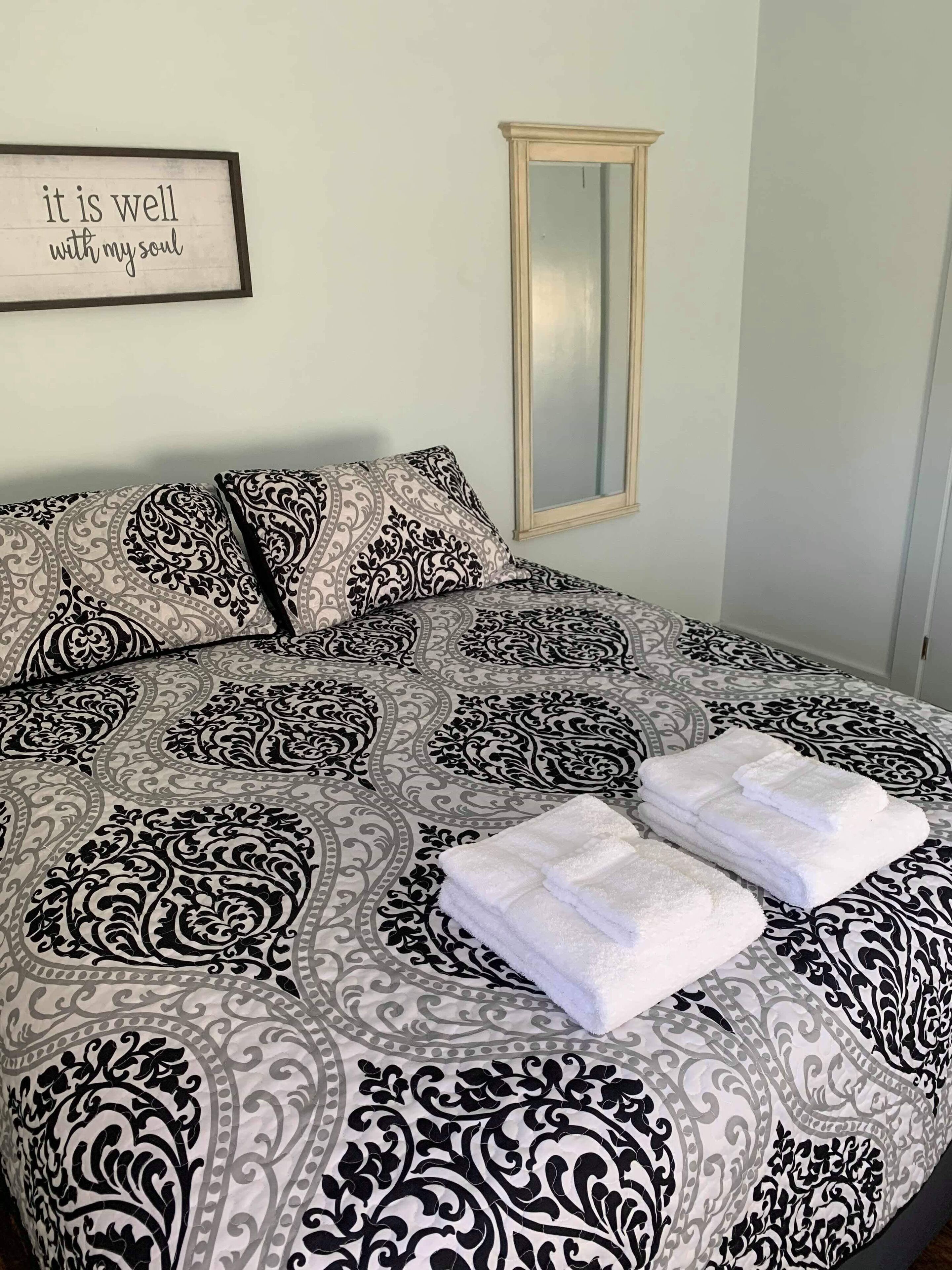 Un lit bien fait, avec une couvre-lit à motifs, deux oreillers et des serviettes blanches pliées. Une citation encadrée est visible au mur.