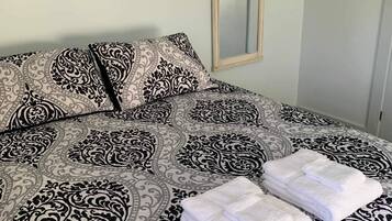 Un lit bien fait, avec une couvre-lit à motifs, deux oreillers et des serviettes blanches pliées. Une citation encadrée est visible au mur.