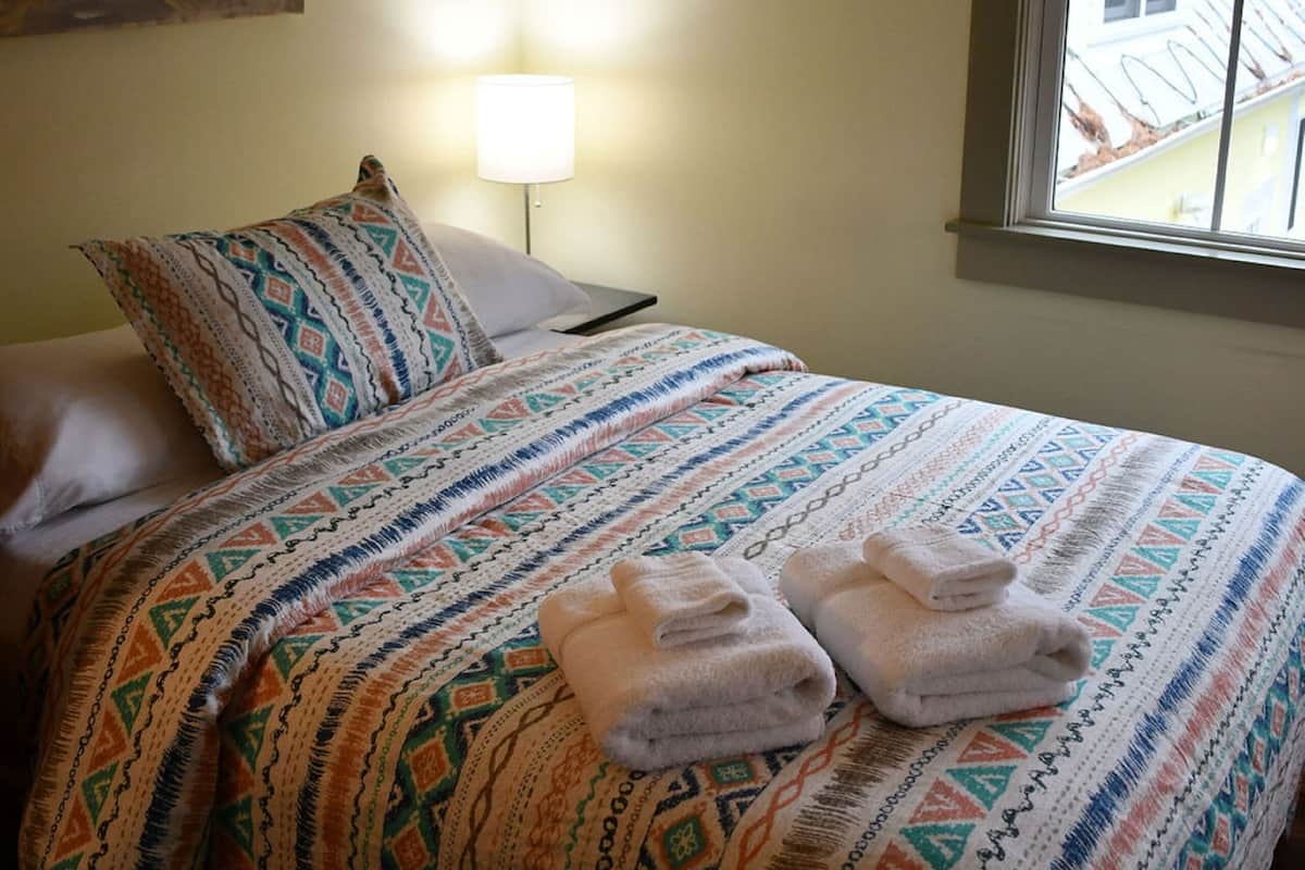 A neatly made bed with patterned bedding and two folded towels, a bedside lamp, and a window with a view of buildings outside.