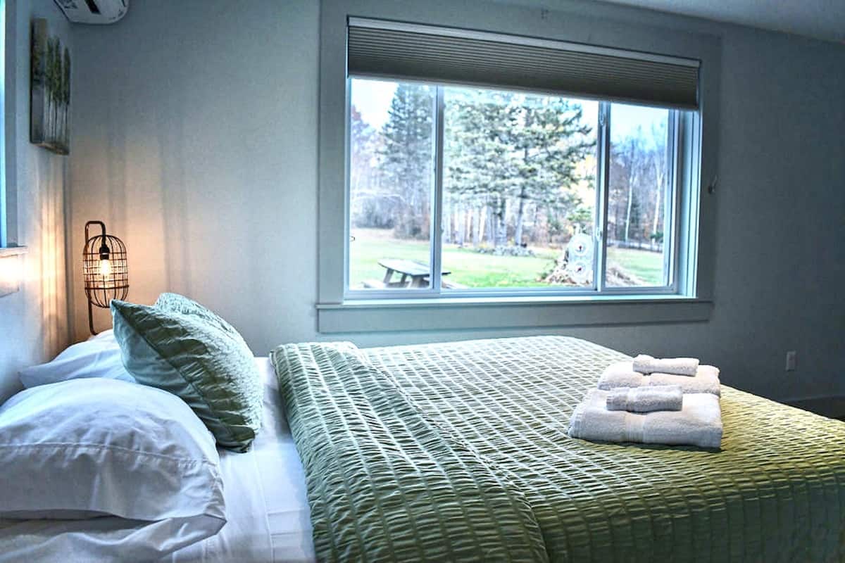 A bedroom with a large window, a bed with green bedding, a bedside lamp, and a view of a yard.