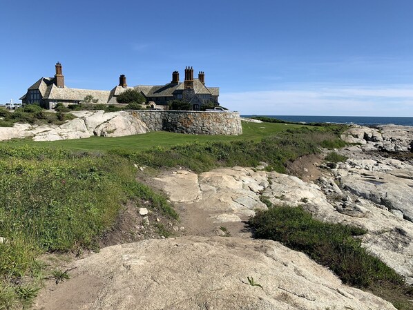 A stone building with multiple chimneys, surrounded by a stone wall and a grassy area with a rocky foreground.