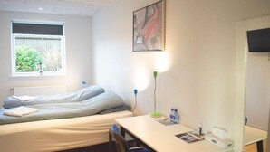 Un dormitorio con cama, escritorio, silla, ventana con vista a follaje y un radiador, además de un cuadro en la pared.