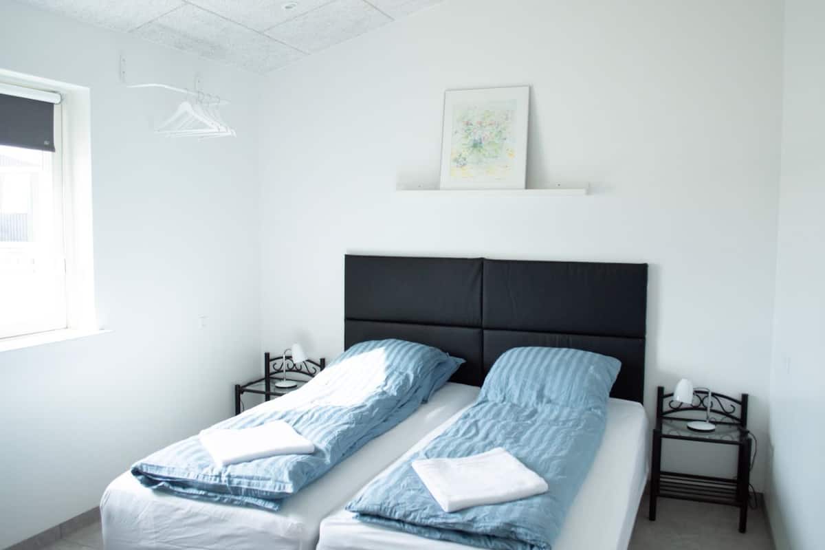 A twin room with two beds, a nightstand, a wall-mounted shelf, and a window with a blackout blind.