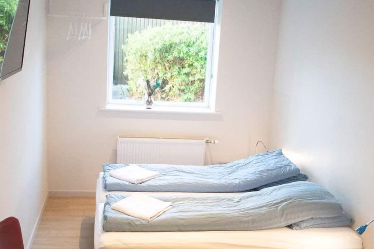 A small, well-lit room with a single bed, a desk, and a chair. A window provides natural light and a view of greenery outside.
