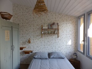 Quarto com cama, prateleira de madeira com livros e objetos decorativos, luminária pendente e janela com cortinas.