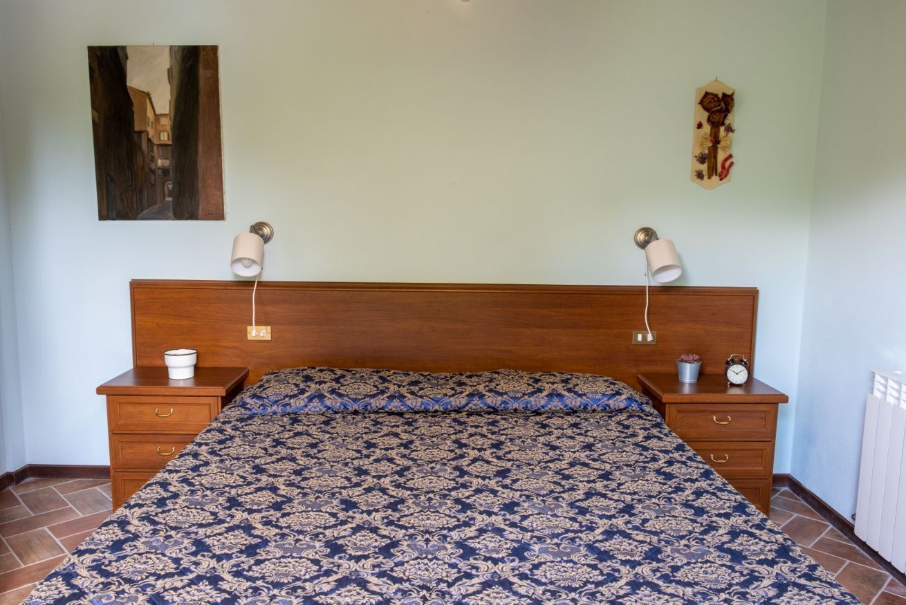 A bedroom with a wooden headboard, a patterned bedspread, bedside tables with lamps, a small clock, and a painting on the wall.