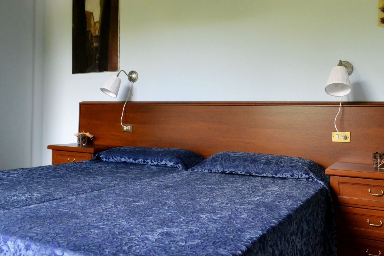 Ein ordentlich bezogenes Bett mit einer blauen, gemusterten Tagesdecke, zwei Nachttischlampen, einem hölzernen Kopfteil und einem kleinen Nachttisch mit Lampe und einem dekorativen Gegenstand.