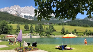 Terraza junto al lago con mesa, sillas y sombrillas, con vistas a una cadena montañosa.