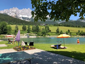 Ein Seeufer mit einer Terrasse, einem Tisch, Stühlen und Sonnenschirmen, mit Blick auf ein Gebirge.