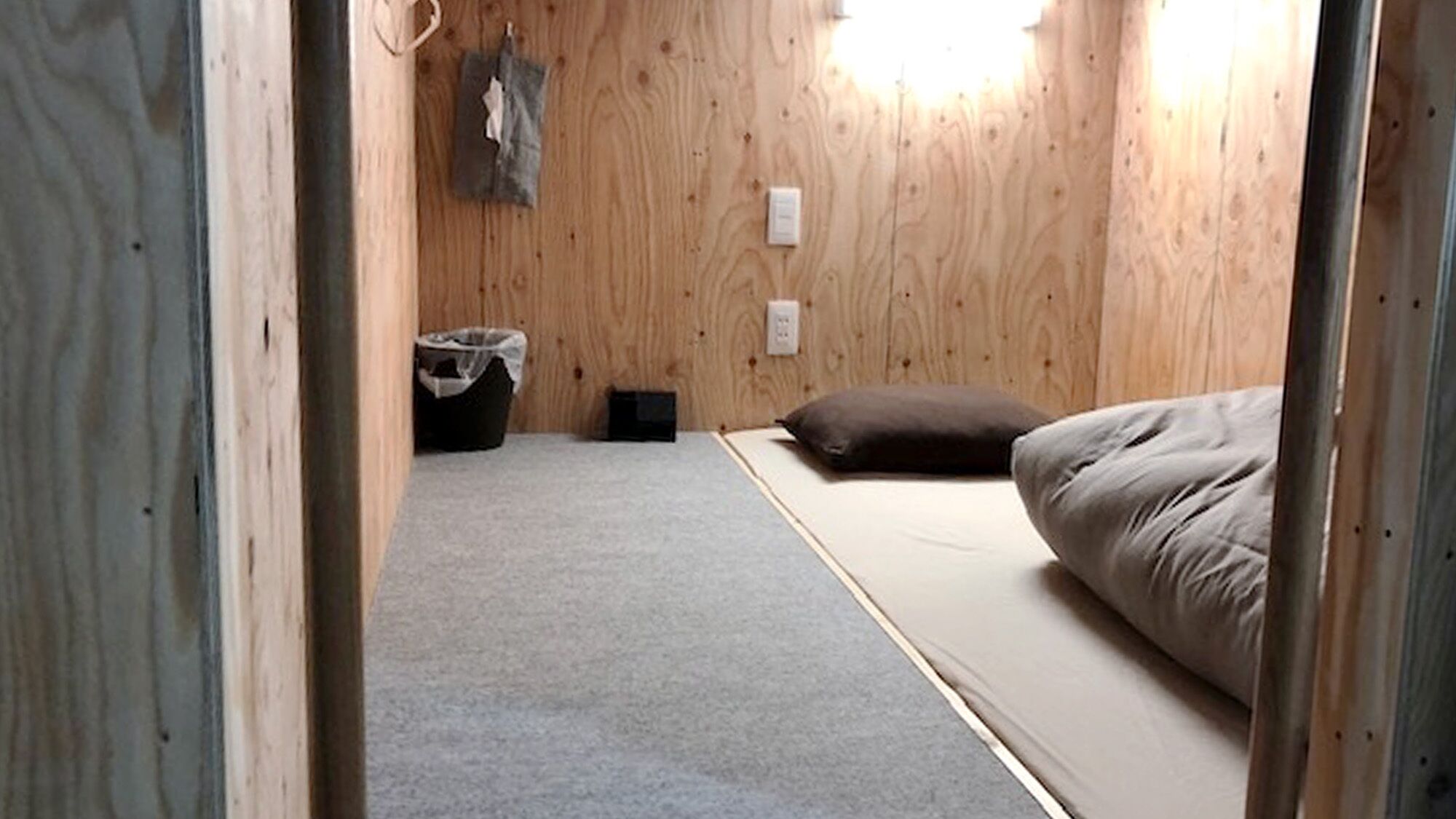 Una habitación con una cama, un pequeño cubo de basura y un enchufe en la pared.