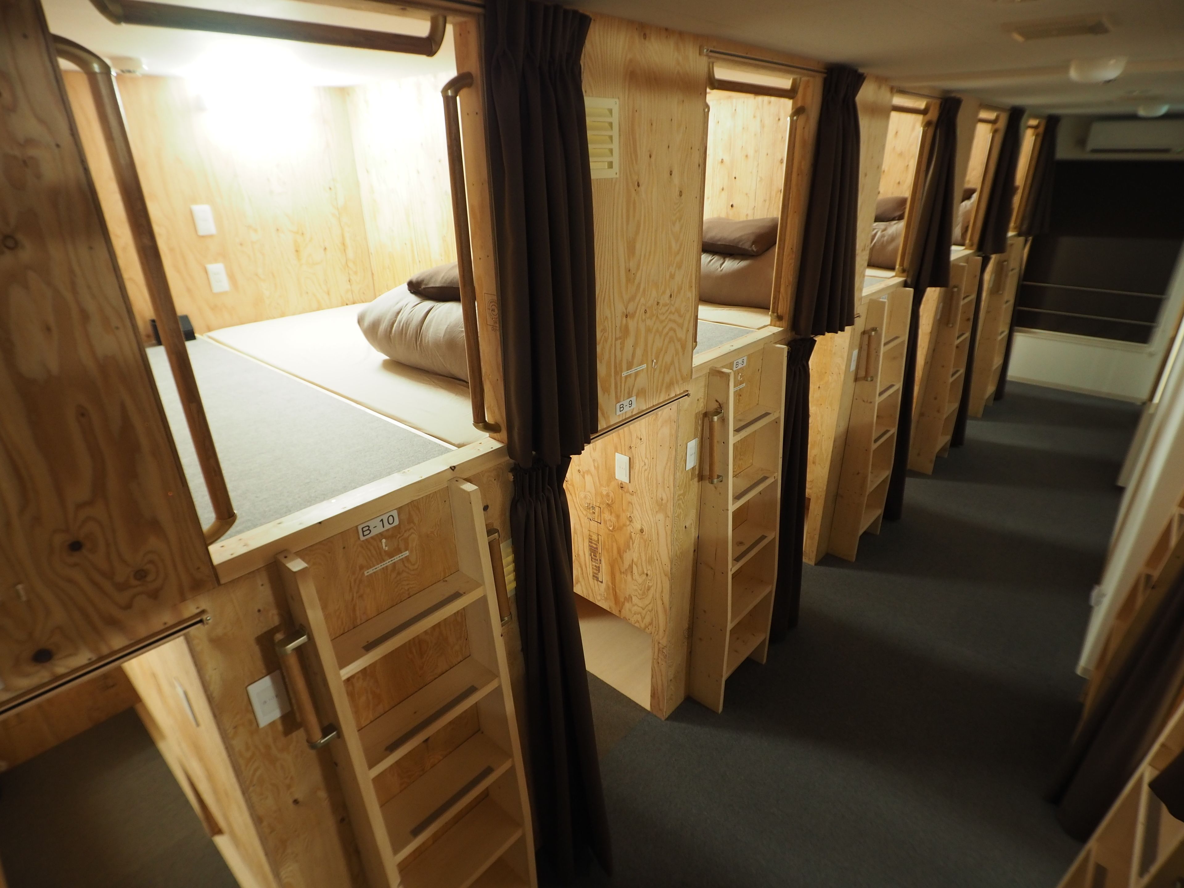 A row of wooden bunk beds with storage drawers, a small table, and a window with curtains.