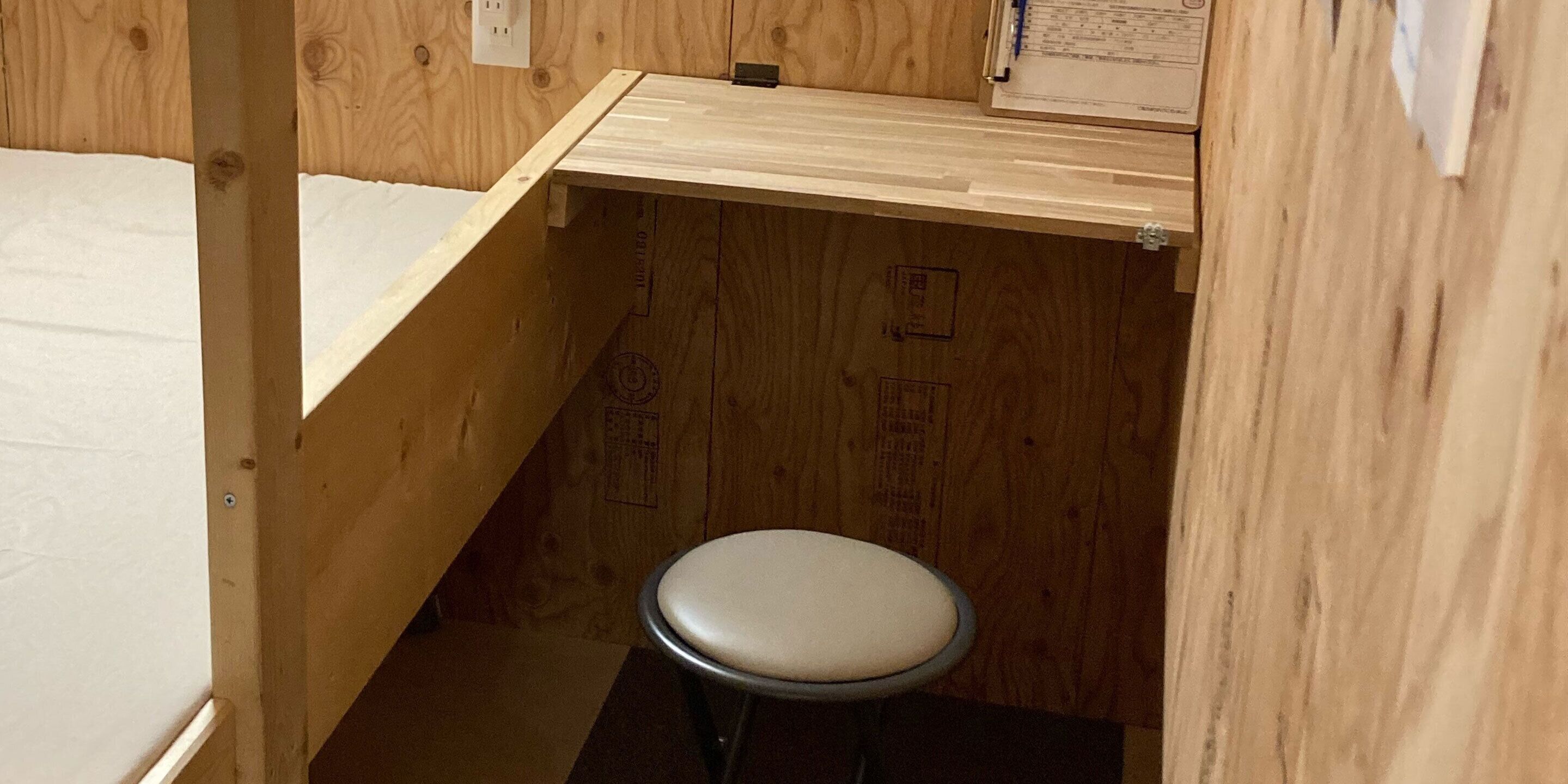 A small wooden room with a single stool, a wooden shelf, and a white mattress.