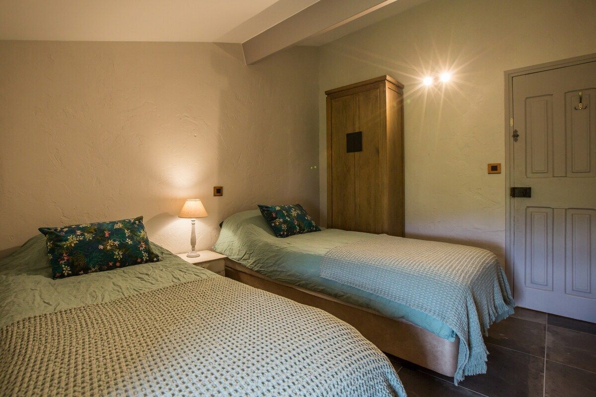A bedroom with two single beds, a wooden wardrobe, a nightstand with a lamp, and a door with a light switch.