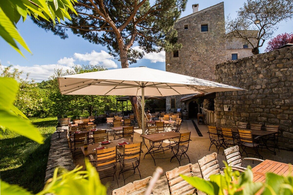 An outdoor dining area with tables and chairs, a large umbrella, and a stone building in the background.