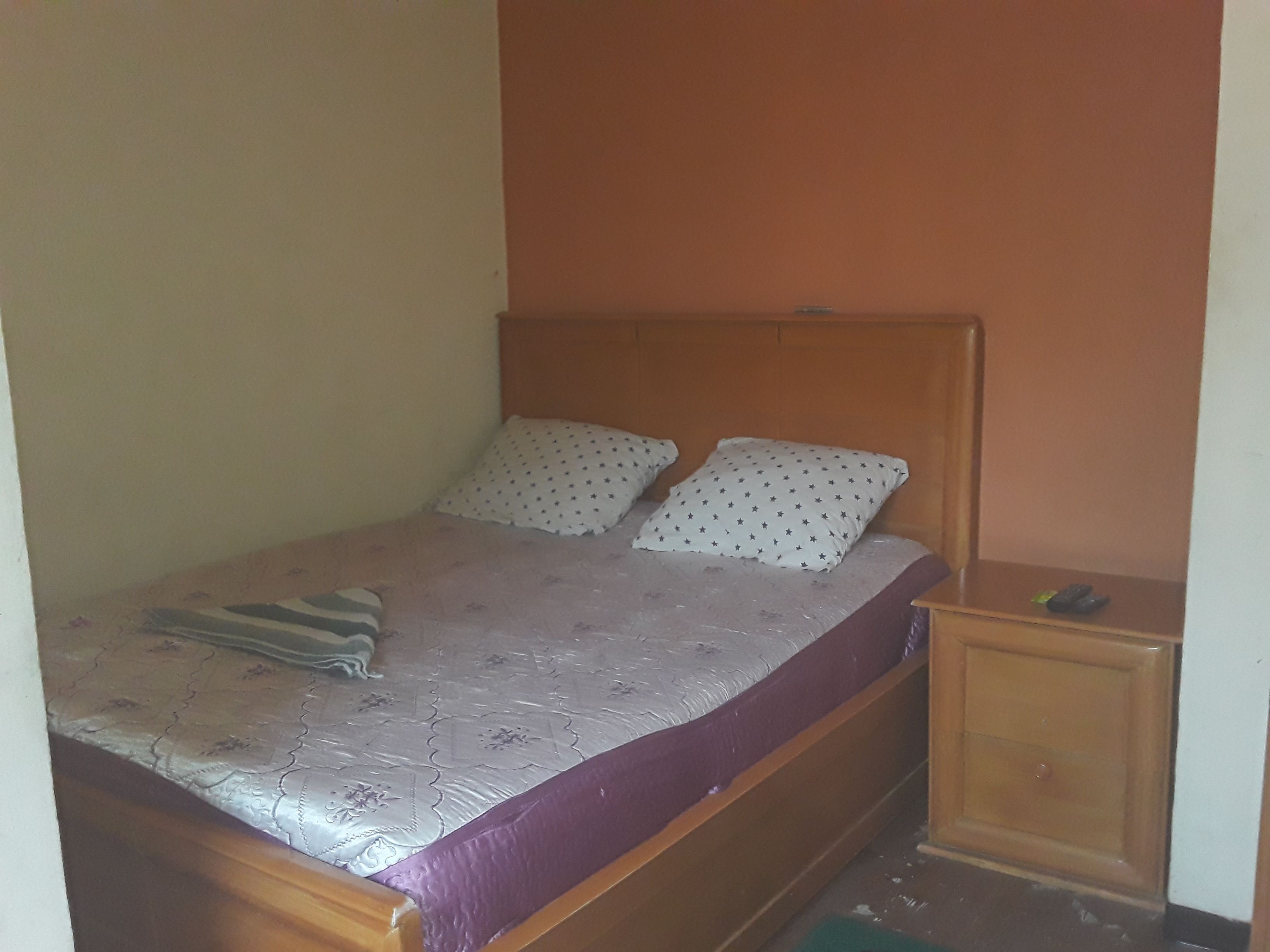 A single bed with a wooden headboard, two pillows with star patterns, and a purple blanket. A small wooden nightstand with a remote control is to the right.