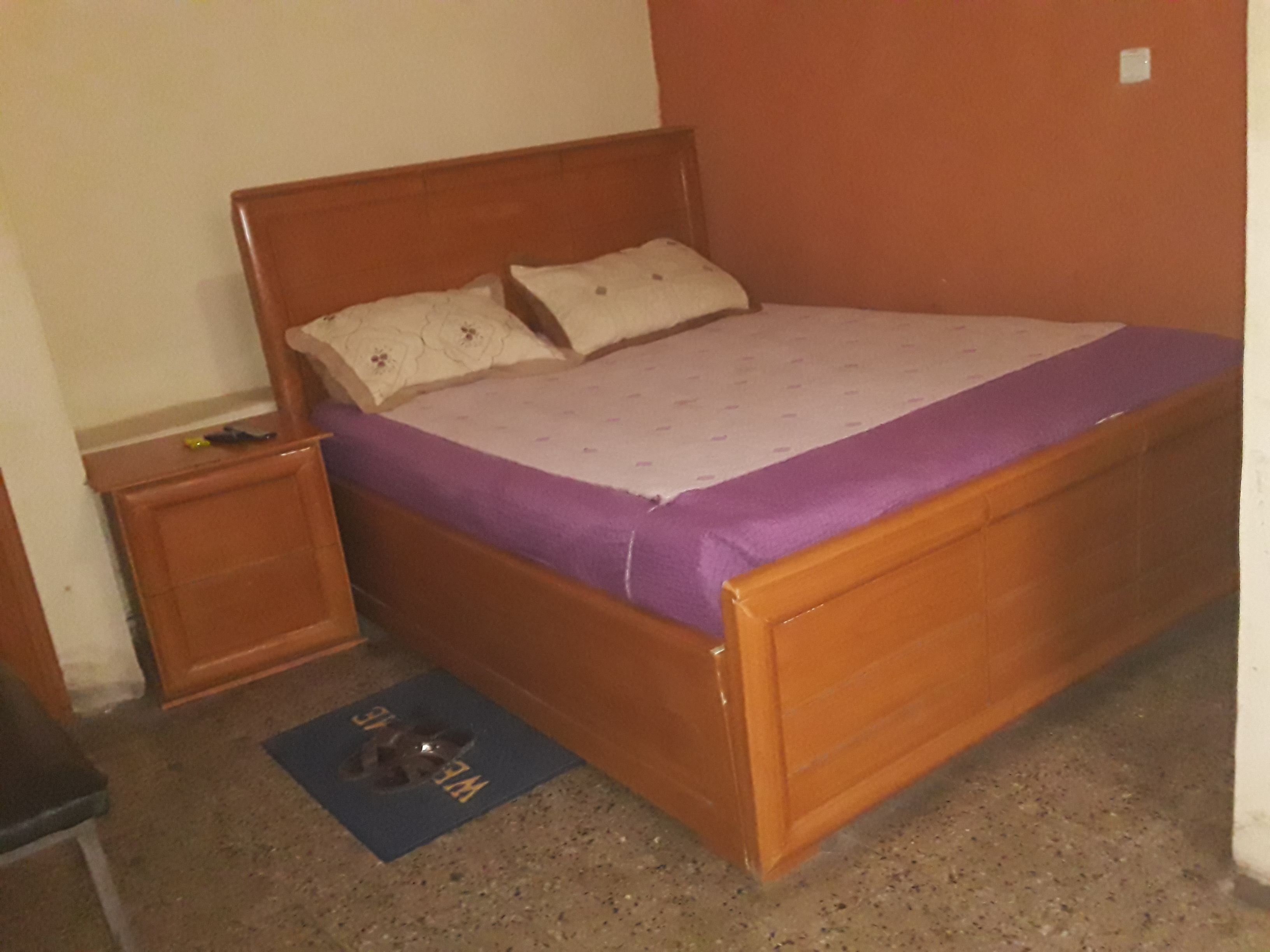 A single-bed room with a wooden headboard, a purple bedspread, and a small bedside table.