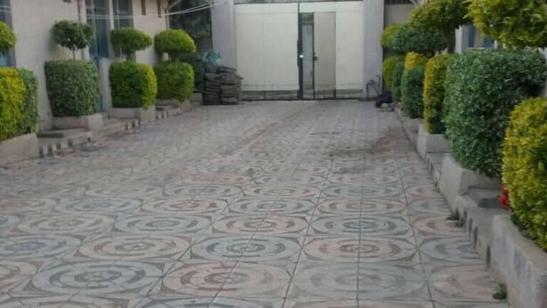 A tiled walkway with a geometric pattern, flanked by neatly trimmed hedges and a white building in the background.