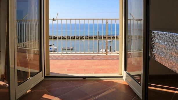 Uma porta aberta dá para uma varanda com vista para o mar e barcos.