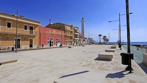 Um calçadão costeiro com céu claro, destacando um farol imponente, uma fileira de edifícios coloridos e um passeio pavimentado com bancos.