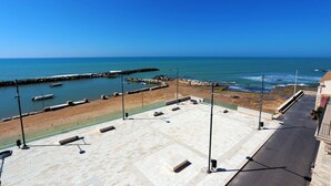 Vista costeira com um calçadão pavimentado, um molhe rochoso e barcos na água.