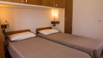 Quarto com duas camas de solteiro, armários de madeira na parede superior e luminárias fixadas na parede.