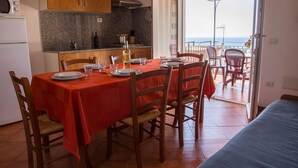 Un comedor con mantel rojo, sillas de madera y vista al mar a través de un ventanal abierto.