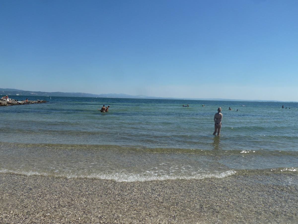 Une plage à l’eau claire, avec un rivage de galets et des montagnes qui se dessinent à l’horizon.