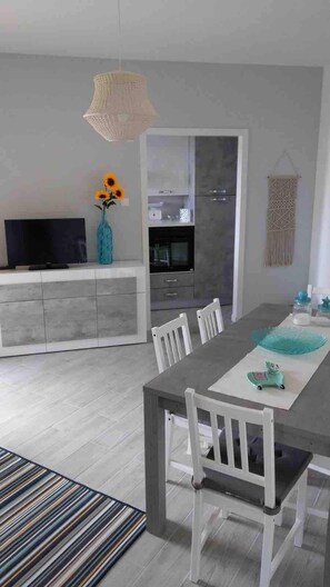 A modern dining area with a long table, white chairs, a television, and a vase with sunflowers. - La Finestra sul mare (Marina Julia, Monfalcone,Italy)