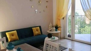 Ein Wohnzimmer mit einem blauen Sofa, einem runden Tisch mit Stühlen, einer dekorativen Wanduhr und einem großen Fenster mit Vorhängen.