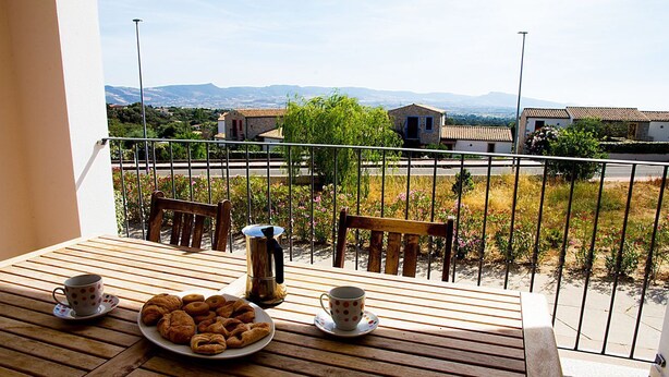 Een balkon met een houten tafel, twee stoelen, een bord gebak en twee kopjes.