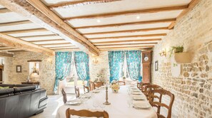 Comedor con una larga mesa dispuesta para una comida, paredes de piedra, vigas de madera y un reloj.