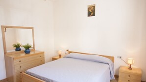 A bedroom with a wooden bed, a nightstand with a lamp, a dresser with a mirror, and two potted plants. - APPARTAMENTO LA PILA - APPARTAMENTO LA PILA (Campo nell'Elba)