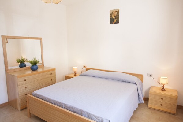 A bedroom with a wooden bed, a nightstand with a lamp, a dresser with a mirror, and two potted plants. - APPARTAMENTO LA PILA - APPARTAMENTO LA PILA (Campo nell'Elba)