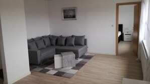 Un salon moderne avec un canapé d’angle gris, un petit pouf blanc et un tapis à carreaux. Un tableau encadré est accroché au mur, et une porte donne sur une chambre à coucher équipée d’un lit et d’une table de chevet.