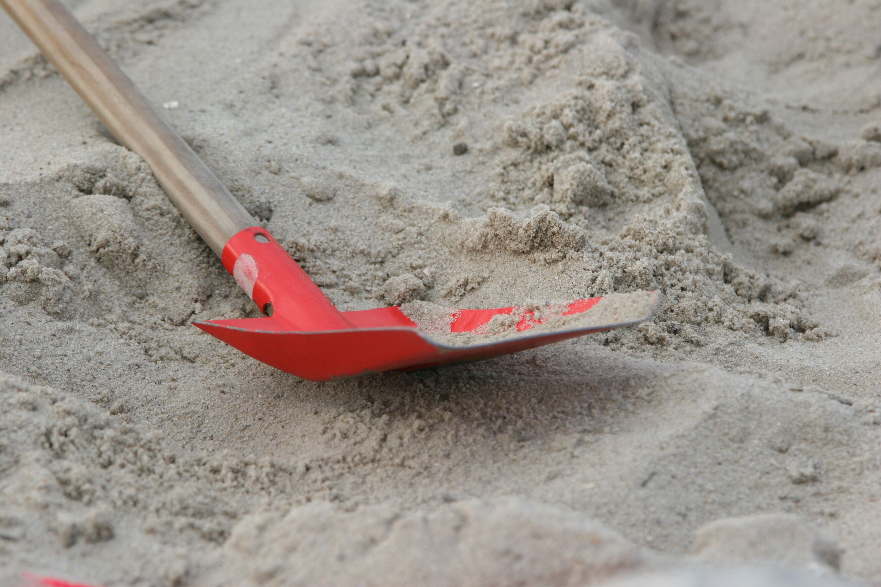 Eine Schaufel mit rotem Stiel und Schneide, teilweise im Sand vergraben.