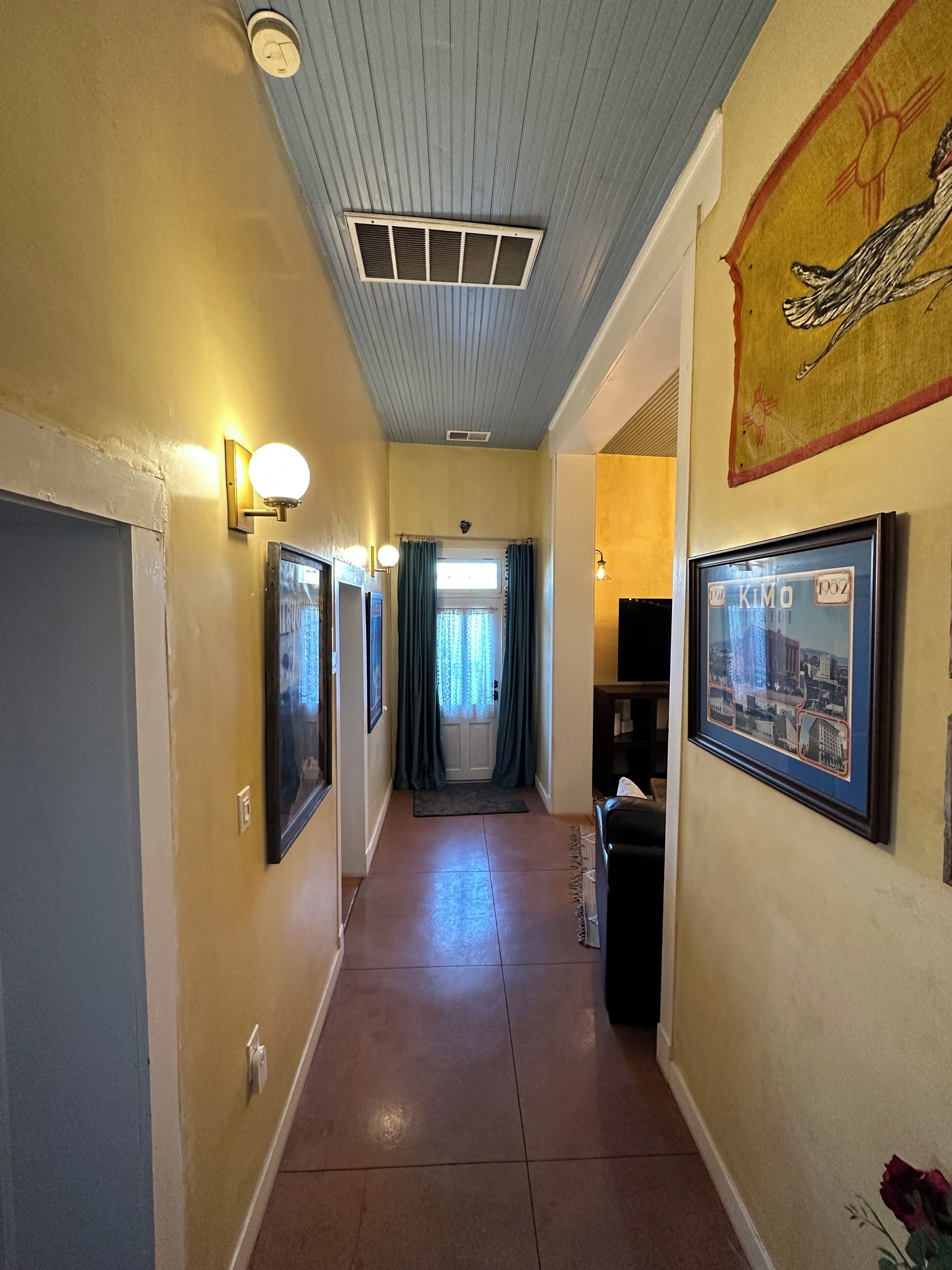 A hallway with a tiled floor, framed pictures on the walls, a ceiling vent, and a door at the end.