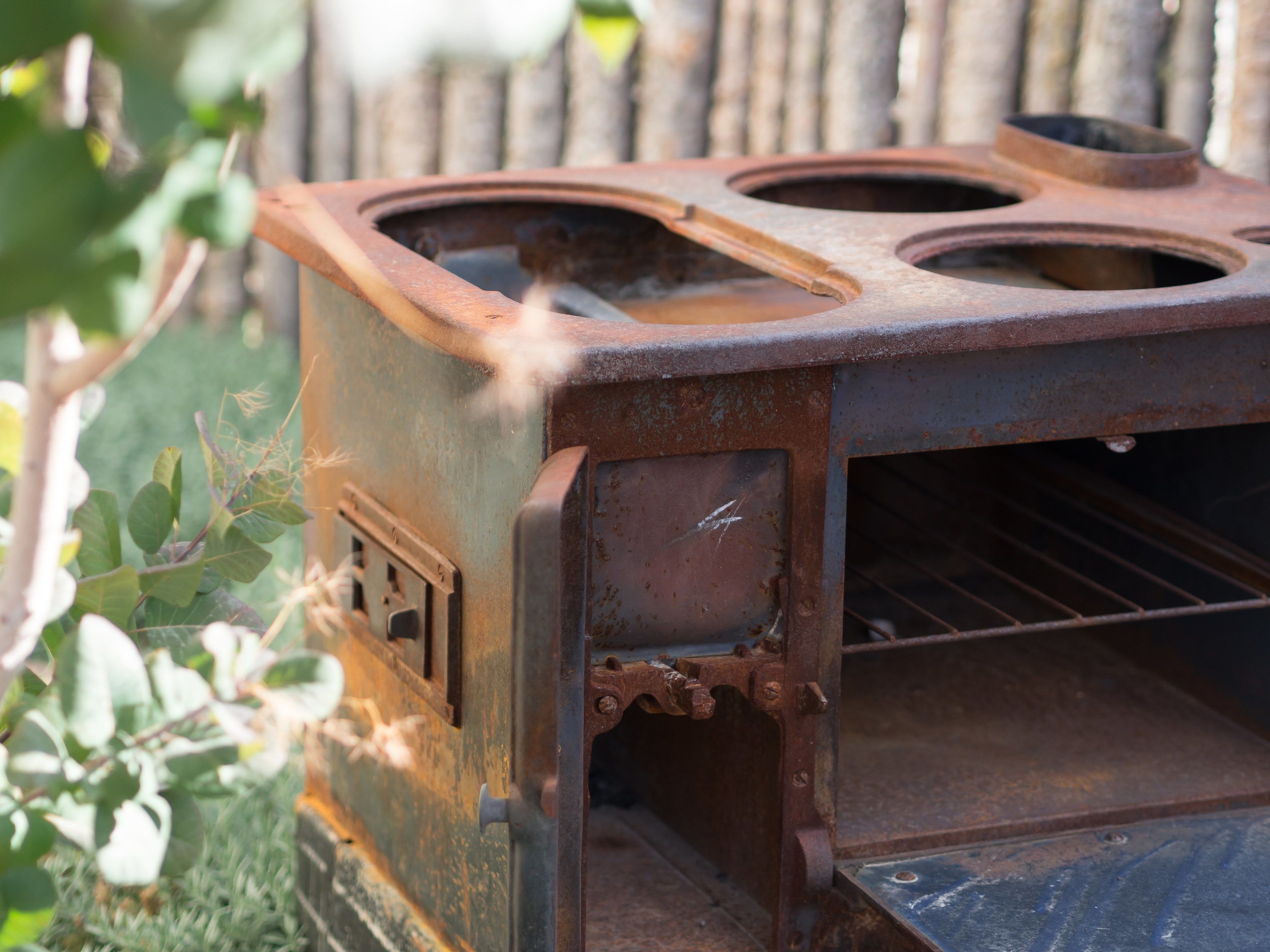 A vintage, rusted stove with four burners and a small vent, set outdoors with greenery in the background.