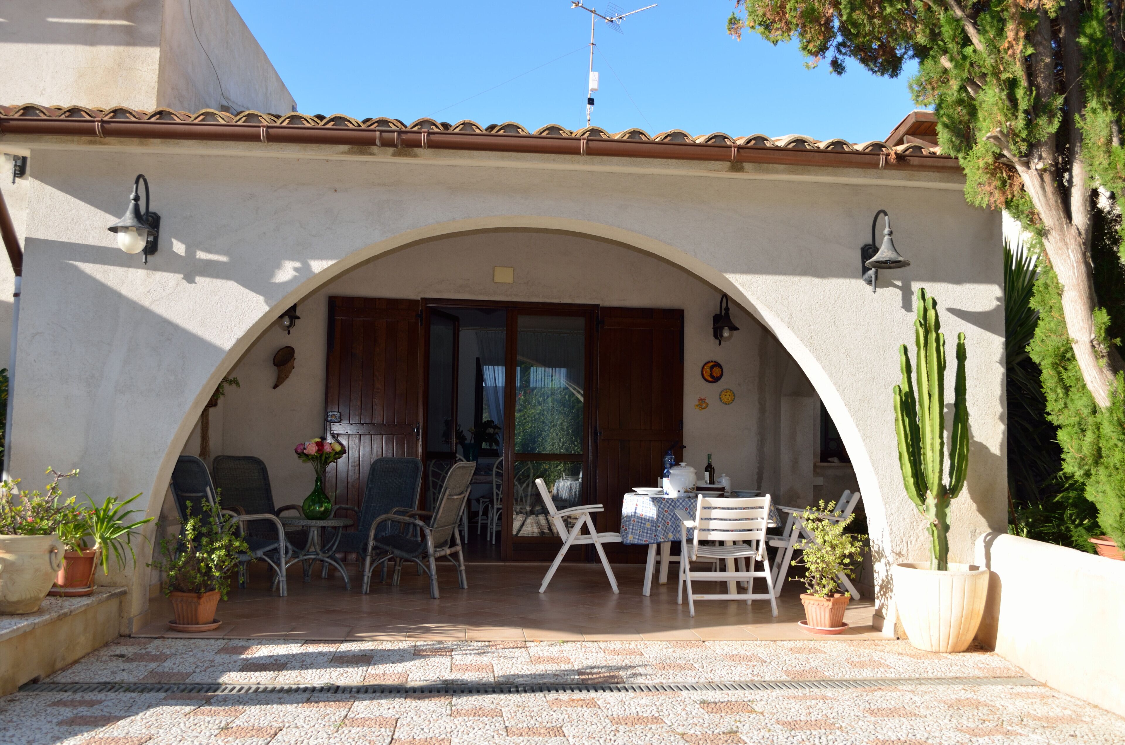 Ein Terrassenbereich im Freien mit Fliesenboden, mehreren Stühlen, einem Tisch und Blumentöpfen.