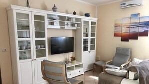 Ein Wohnzimmer mit Fernseher, weißen Schränken, zwei Sesseln und einem Sofa.