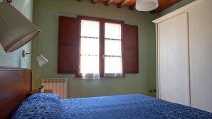 Un dormitorio con una cama, una ventana con contraventanas de madera, un radiador y una lámpara blanca.