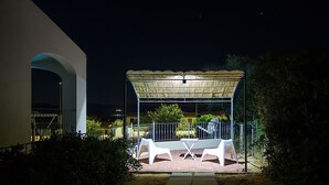En udendørs patio med et overdækket siddeområde, en sti og en bygning i baggrunden.