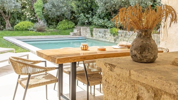 Ein Essbereich im Freien mit Pool, Steintisch, Korbstühle und einer Vase mit Trockenpflanzen.